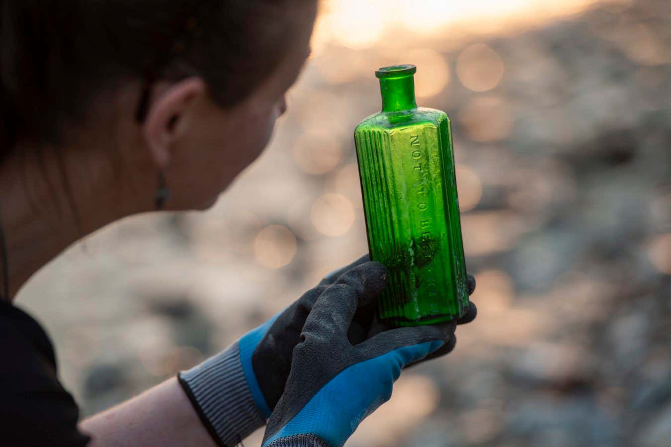Mudlark in gloves examines an old, green glass poison bottle with embossed text ? London Museum Mudlarkers photographed on the Thames between Millenium Bridge and Southwark Bridge along with finds, landscape views and close ups.